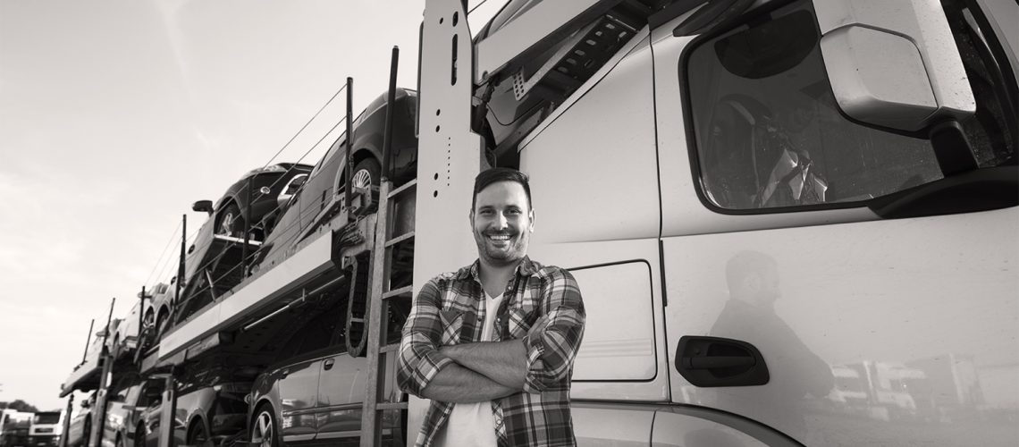 Portrait of professional smiling truck driver with crossed arms transporting cars to the market.